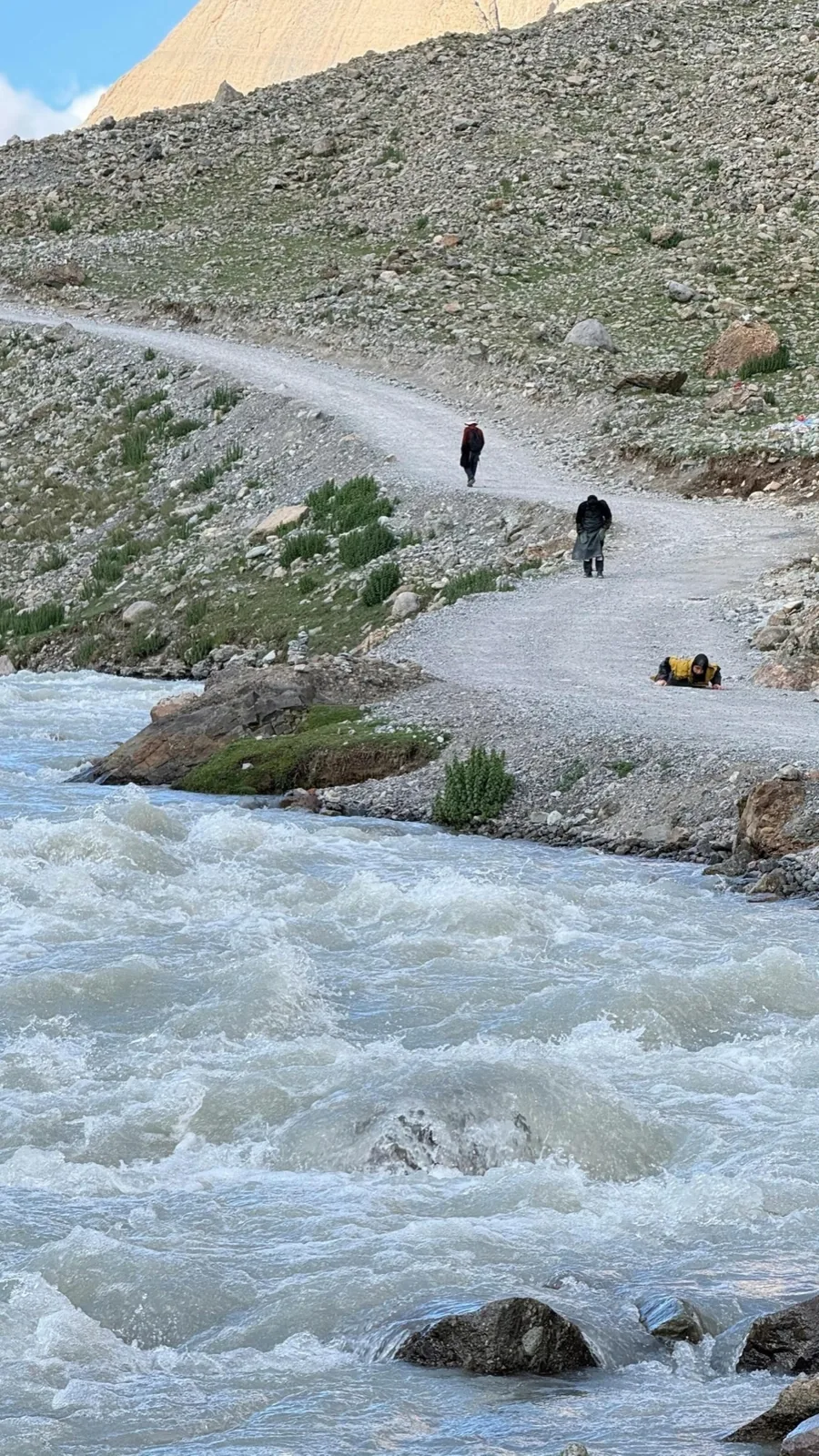 Small figures walking along a pale river path below a rocky slope